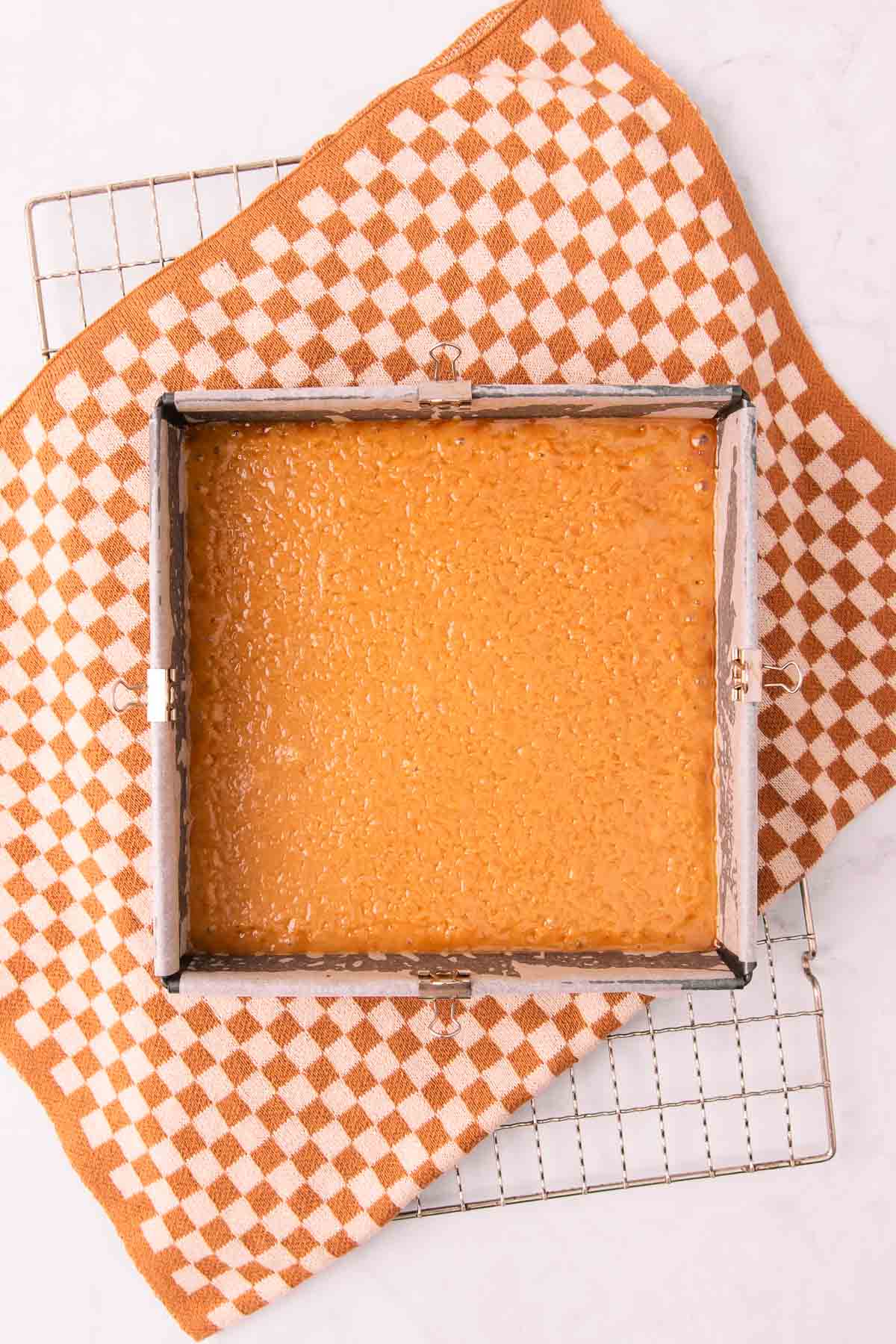 The baked caramel layer in a square pan on a cooling rack with a brown and tan checked tea towel underneath.