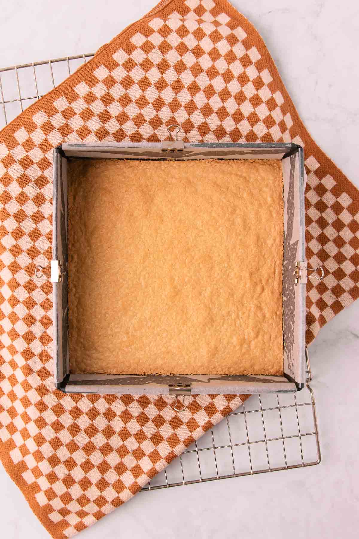 The golden brown baked slice base in a square pan on a cooling rack with a brown and tan checked tea towel underneath.