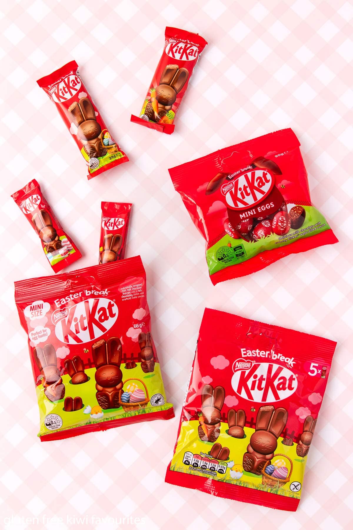 A packet of KitKati mini eggs, two larger Kitkat bunnies, and two packets of Kitkat mini bunnies, with one packed open and two mini bunnies sticking out, on a pink and white checked background.
