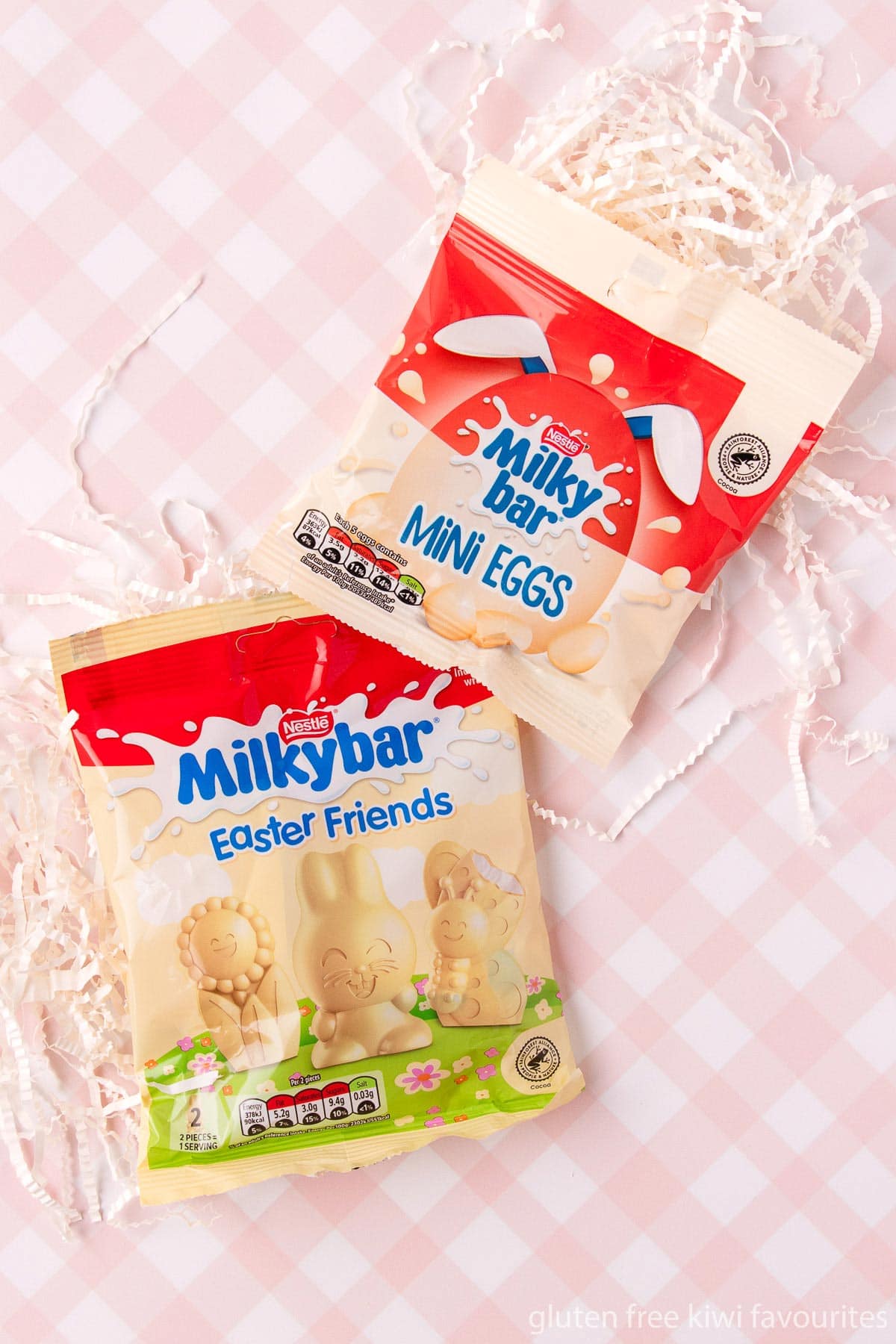 A bag of Milky Bar mini eggs and Easter Friends on top of some white shredded paper on a pink and white checked background.