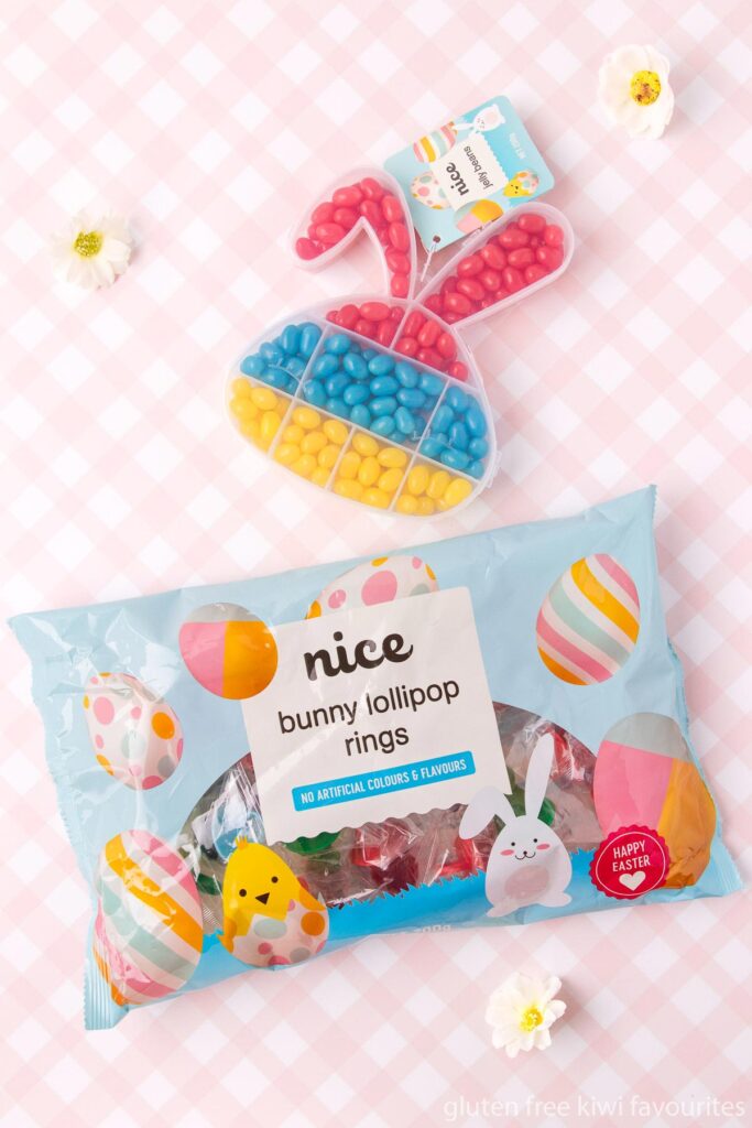 A bag of Nice brand bunny lollipop rings, and a bunny head shaped plastic container of red, blue and yellow jelly beans on a pink and white checked background.