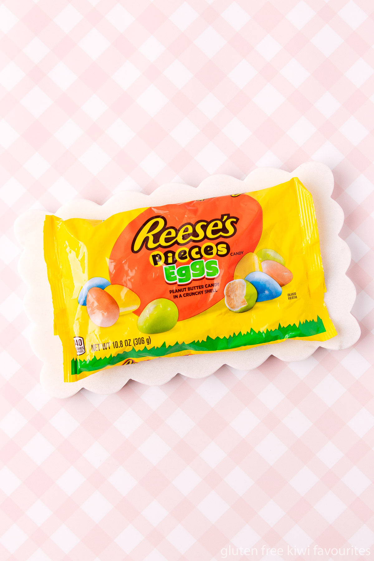 A bright yellow bag of Reese's Pieces Easter Eggs, on a scalloped-edge marble tray, on a pink and white checked background.