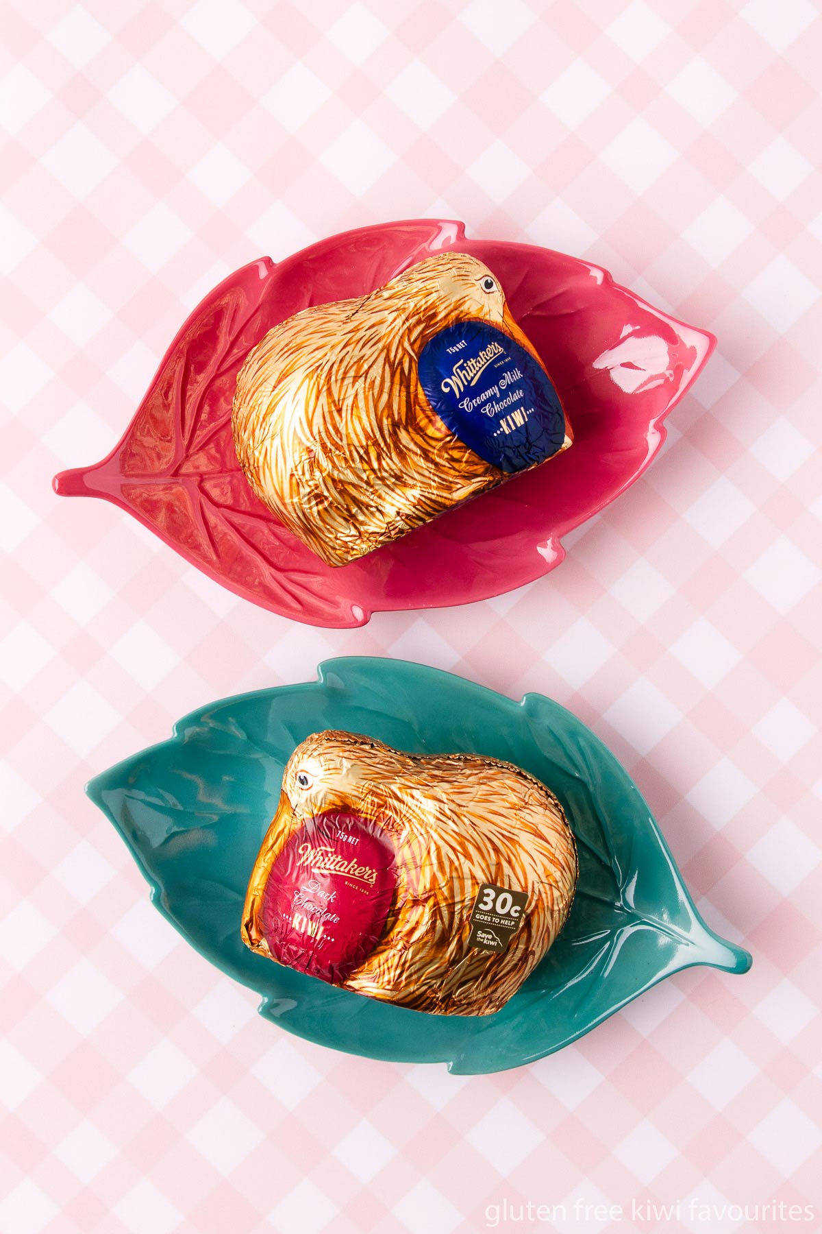 Two Whittakers chocolate kiwis - one milk chocolate and one dark chocolate, on leaf-shaped plates on a pink and white checked background.