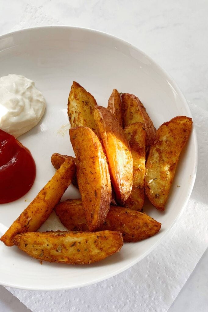 A white plate of potato wedges with a crispy coating, with tomato ketchup and sour cream on the side.