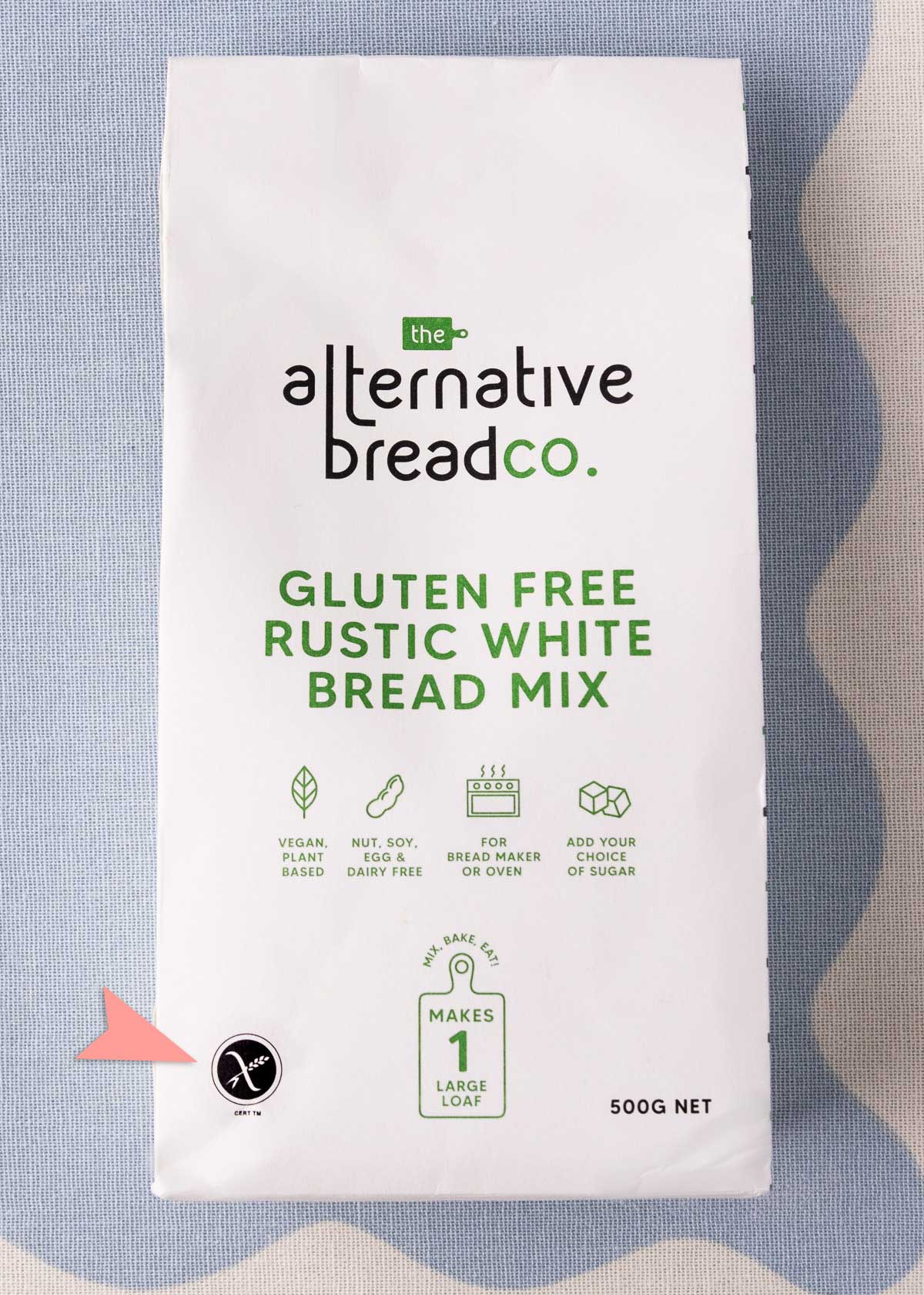 A bag of The Alternative Bread Co. gluten free rustic white bread mix which is a white paper bag with black and green writing, and there is a pink arrow pointing to the Crossed Grain Logo, a round circle with a stalk of wheat crossed through with a line.